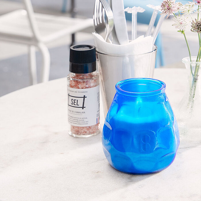 Ein einzelnes, brennendes Bolsius Windlicht in einem orangefarbenen Glasgefäß steht auf einem rustikalen Holztisch. Im Hintergrund sind ein Teller mit Besteck und Servietten, sowie eine Vase und eine Pfeffermühle zu sehen.