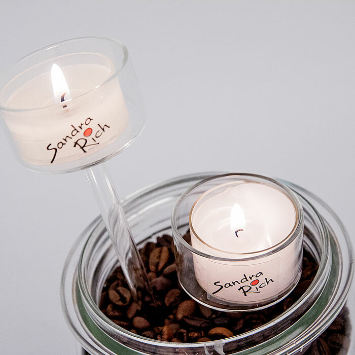 Zwei brennende Teelichter auf hohen Glasständern in einer quadratischen Glasvase, die zur Hälfte mit kleinen weißen Steinen gefüllt ist. Drumherum und innerhalb der Vase befinden sich dekorative, rosa-farbene Drahtkugeln. Die Glasvase steht auf einem rote