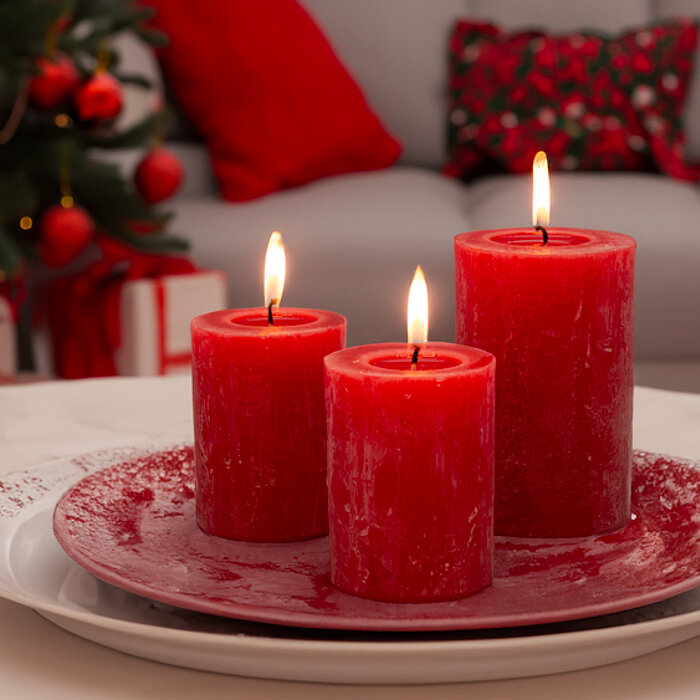 Vier brennende DecoLite Stumpenkerzen in den Farben Weiß und Beige stehen auf einem runden Tablett auf einem hellgrauen Tisch. Im Hintergrund befindet sich ein modernes Wohnzimmer mit einem beige-grauen Sofa, das mit grauen Kissen dekoriert ist, und einem