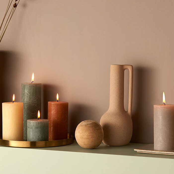 Sechs brennende Bolsius Rustikkerzen in verschiedenen Größen und Farben, darunter Weiß, Grün und Rosa, stehen auf einer Holzablage. Daneben befinden sich eine rosa Blume in einer kleinen Vase und ein Set aus Keramikgeschirr. Die Szene wirkt warm und einla