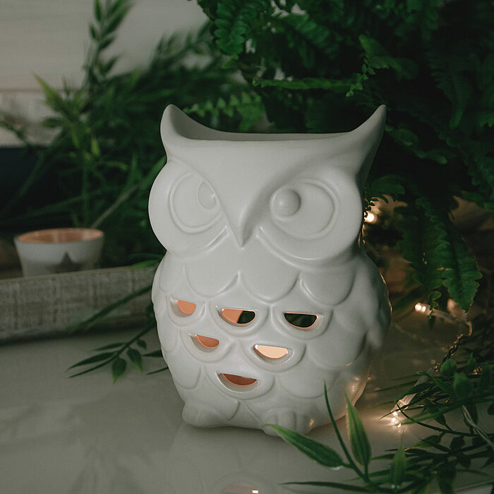 Eine weiße Eulenkerze mit durchbrochenen Mustern steht auf einem Tisch, umgeben von kleinen leuchtenden Lichterketten und grünen Pflanzen. Im Hintergrund ist ein weißer, dekorierter Blumentopf zu sehen. Die Stimmung des Bildes ist ruhig und gemütlich.