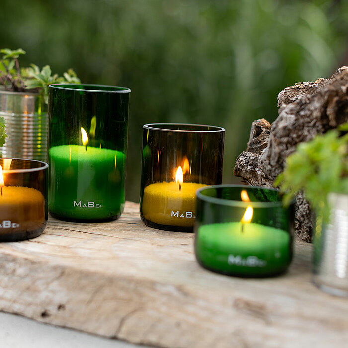 Vier brennende Kerzen des Herstellers Wenzel in grünen und braunen Glasbehältern sind auf einem rustikalen Holztisch platziert. Zwischen den Kerzen befinden sich kleine Pflanztöpfe aus Metall mit grünen Pflanzen. Im Hintergrund sind verschwommene grüne Pf