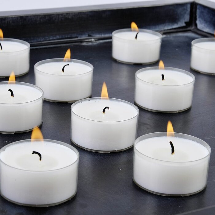 Zwölf brennende weiße Teelichter des Herstellers Müller sind auf einem schwarzen Tablett angeordnet. Die flackernden Flammen verbreiten ein gemütliches Licht.