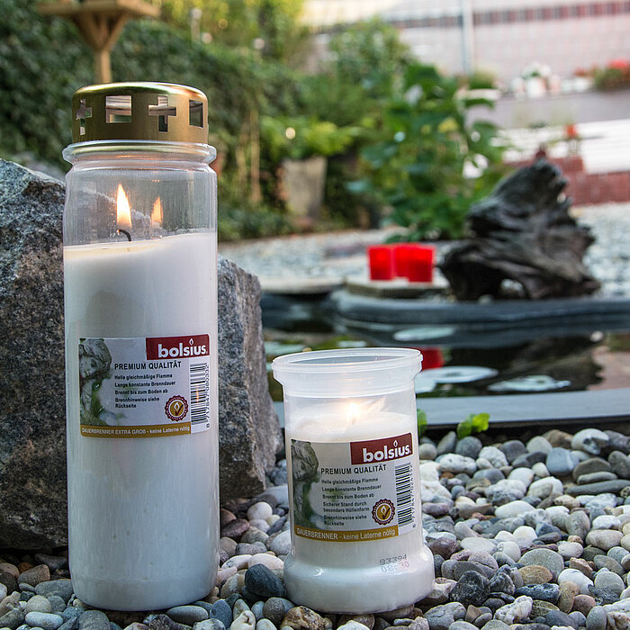 Zwei brennende Bolsius Grabkerzen, eine größere und eine kleinere, stehen auf einem Kiesbett vor einem dekorativen Teich. Im Hintergrund sind Pflanzen und ein großer Felsen zu sehen. Die größere Grabkerze hat einen goldfarbenen Deckel.