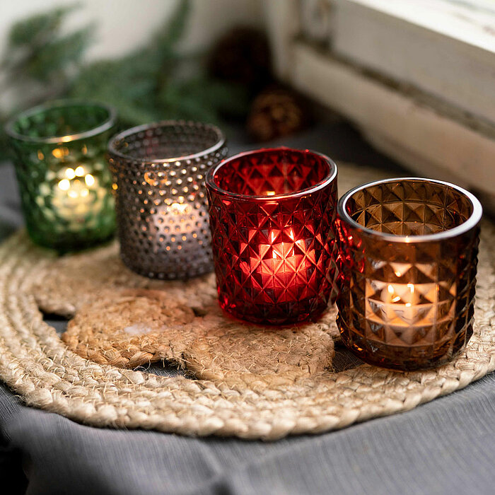Vier kleine, leuchtende Teelichter des Herstellers Sandra Rich stehen in dekorativen, geometrisch gemusterten Glashaltern in den Farben Grün, Grau, Rot und Braun auf einer runden, geflochtenen Unterlage. Im Hintergrund sind verschwommene Zweige und Zapfen