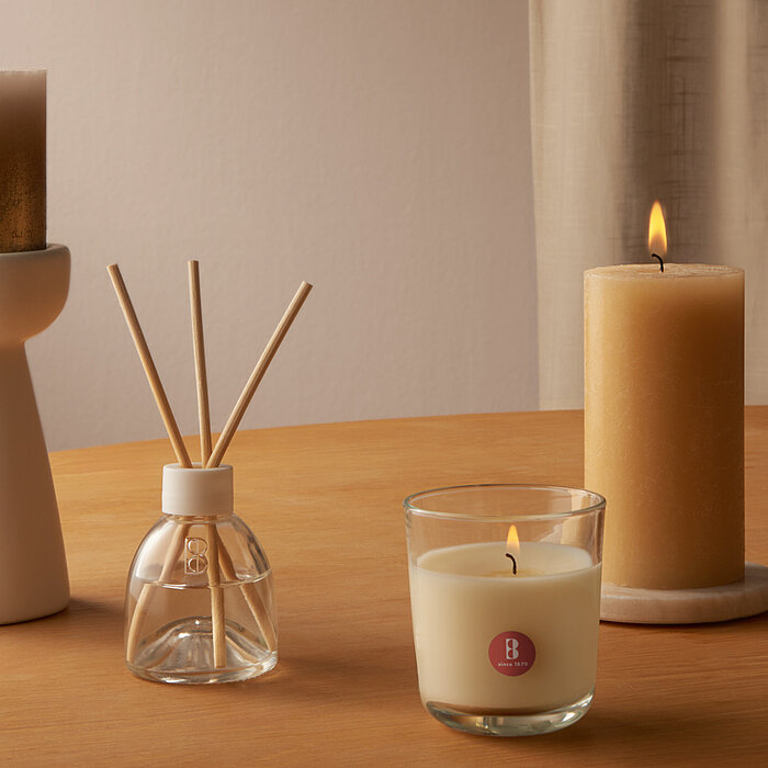 Drei brennende Duftkerzen von Bolsius in verschiedenen Größen und Formen, darunter eine weiße Stumpenkerze, eine rote Stumpenkerze und eine kleine rote Duftkerze in einem Glasgefäß. Die Kerzen stehen auf zwei unterschiedlich großen Tischen, die mit einem 