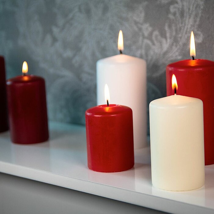 Sechs brennende DecoLite Kerzen in den Farben Rot und Weiß stehen in verschiedenen Größen auf einem weißen Regal vor einer grauen, gemusterten Tapete. Die Stimmung ist ruhig und festlich.