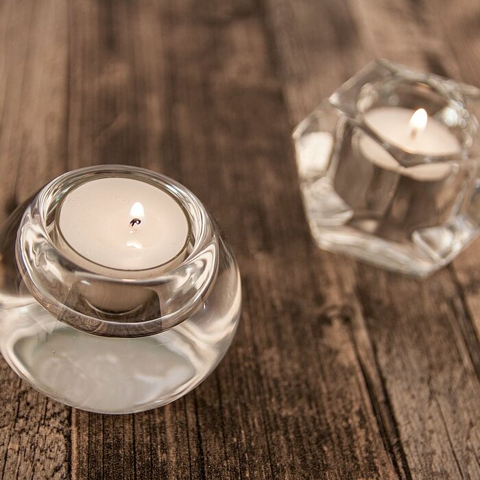 Drei brennende Teelichter stehen in transparenten, geometrisch geformten Glasbehältern auf einer rustikalen Holzoberfläche. Diese eleganten Teelichthalter sind vom Hersteller "Sandra Rich".