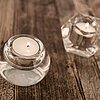 Drei brennende Teelichter stehen in transparenten, geometrisch geformten Glasbehältern auf einer rustikalen Holzoberfläche. Diese eleganten Teelichthalter sind vom Hersteller "Sandra Rich".