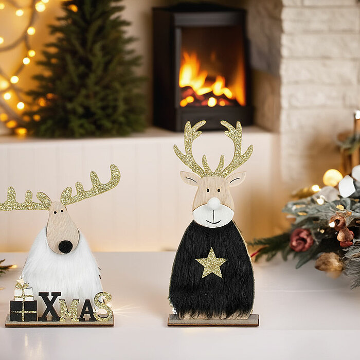 Im Bild sind zwei festliche Dekorationen vom Hersteller „Edelman“ zu sehen: Zwei Rentierfiguren mit Glitzergeweihen, eine in schwarzem und eine in weißem Fell. Beide tragen jeweils ein kleines Geschenk und den Schriftzug „XMAS“. Im Hintergrund leuchten ei