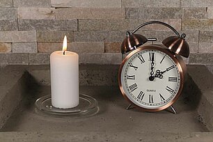 In einer eckigen grauen Betonschale steht eine brennende weisse Bolsius professional Stumpenkerze mit den Maßen 100/48 mm. Rechts neben der Stumpenkerze befindet sich ein schöner alter Wecker in Kupferfarbe, der die Uhrzeit 2 Uhr anzeigt. Im Hintergrund sieht man eine steinerne Mauer.