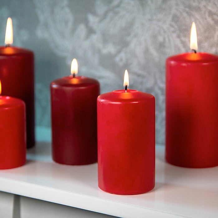 Fünf brennende rote Stumpenkerzen von DecoLite in verschiedenen Größen stehen auf einem weißen Regal. Der Hintergrund zeigt eine graue, floral gemusterte Tapete, die eine elegante Atmosphäre schafft.