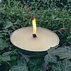 Eine brennende Kerze des Herstellers Wenzel in einer Aluschale, umgeben von üppigem Efeu und grünen Pflanzen im Freien. Die sanfte Flamme schafft eine beruhigende Atmosphäre inmitten der Natur.