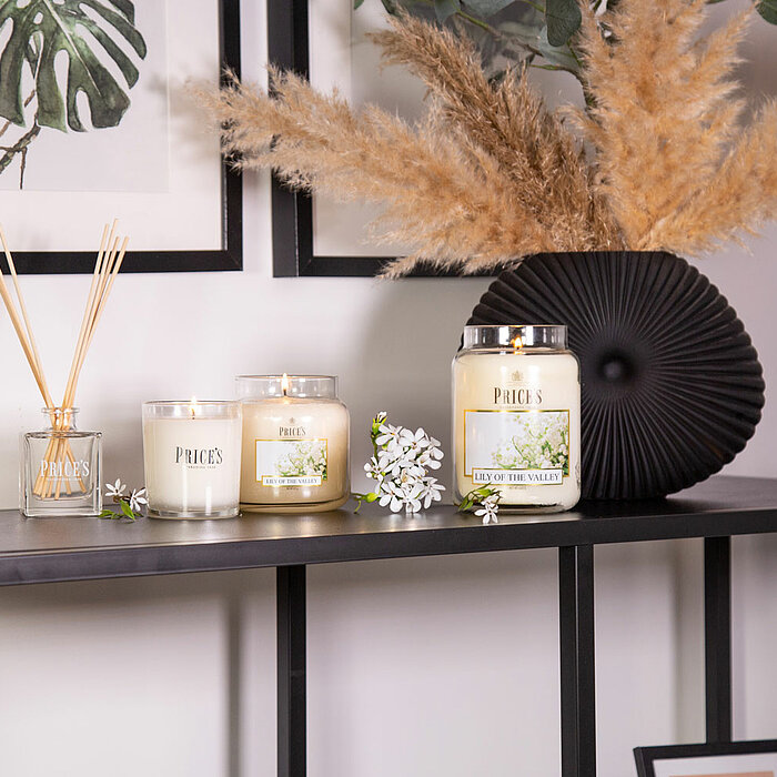 Auf einem schwarzen Metallregal stehen mehrere Duftkerzen von "Prices Candles". Es gibt vier Kerzen in verschiedenen Größen, die in weißen und klaren Gläsern arrangiert sind. Neben den Kerzen ist ein kleiner Glasbehälter mit Duftstäbchen von "Prices Candl