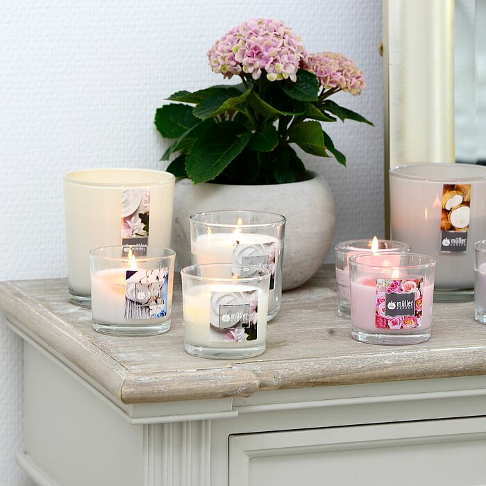 Acht brennende Kerzen vom Hersteller Müller in verschiedenen Designs, einschließlich weißer, rosa und grauer Gläser, stehen auf einer hellen Kommode. Daneben befindet sich eine Vase mit einer rosa Hortensie. Im Hintergrund ist ein Spiegel zu sehen.