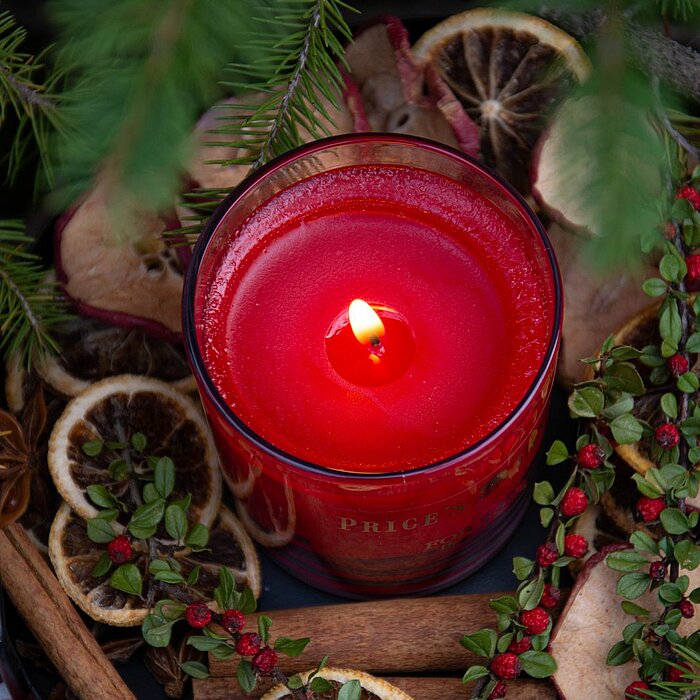 Eine rote, brennende Kerze von Prices Candles befindet sich in einem Glasgefäß und steht auf einem Bett aus getrockneten Apfelscheiben, Zimstangen, Orangenscheiben und grün-rotem Dekor. Im Hintergrund sind grüne Tannenzweige zu sehen, die eine festliche S