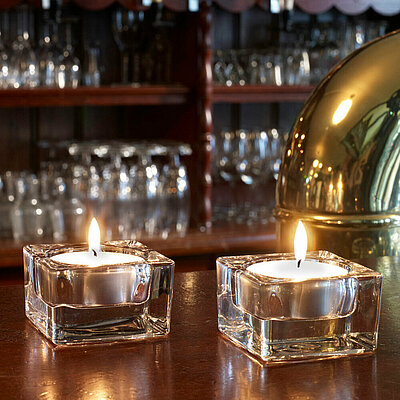 Auf einer Gastronomischen Bar stehen zwei eckige transparente Glas-Teelichthalter mit je einem brennenden weißen Teelicht. Im Hintergrund sieht man mehrere verschiedene Gläser und am rechten Bildrand erkennt man einen goldenen Zapfhahn.