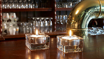 Auf einer Gastronomischen Bar stehen zwei eckige transparente Glas-Teelichthalter mit je einem brennenden weißen Teelicht. Im Hintergrund sieht man mehrere verschiedene Gläser und am rechten Bildrand erkennt man einen goldenen Zapfhahn.