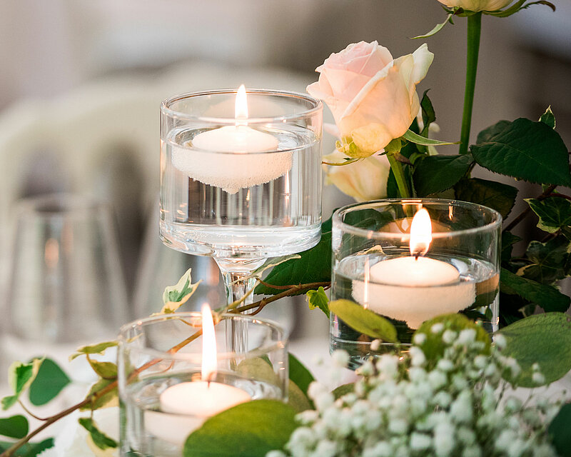 Das gesamte Bild strahlt eine festliche Atmosphäre aus, ähnlich dem Charme einer Hochzeit. Man sieht einen eingedeckten Tisch mit leeren Weingläsern und Servietten. Im Vordergrund befinden sich verschiedene Blumenarrangements mit grünen Blättern, weißen Blüten und einigen rosafarbenen Rosen. Zwischen den Arrangements stehen drei unterschiedlich hohe Schwimmkerzenhalter aus Glas, die mit Wasser gefüllt sind. Auf der Wasseroberfläche schwimmt jeweils eine brennende Schwimmkerze, die ein angenehmes Kerzenlicht verbreiten.