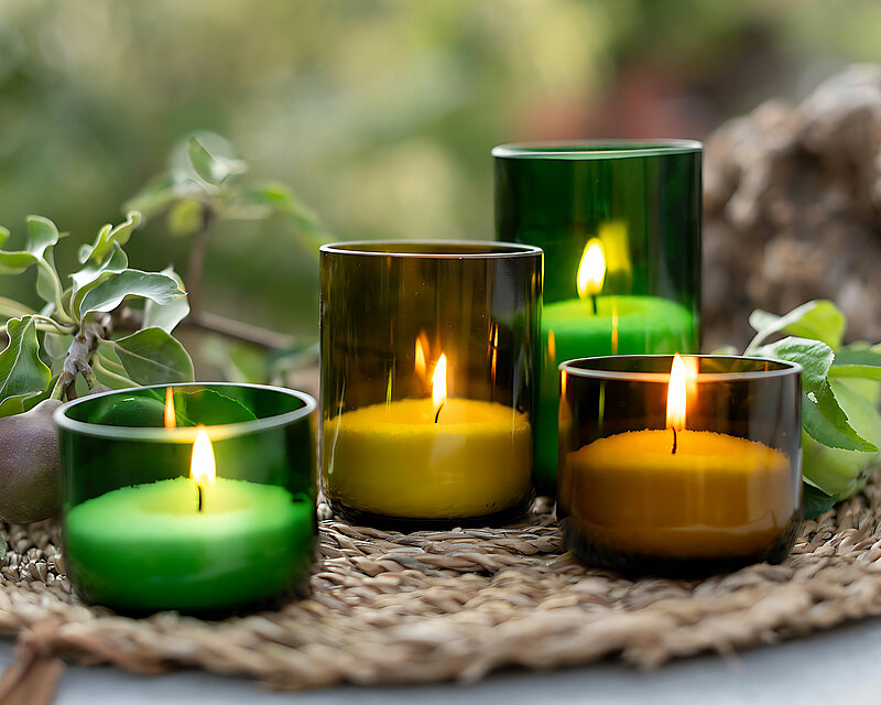 Mehrere grüne und gelbe Kerzengläser gefüllt mit dem weissen Wenzel Wachsgranulat stehen auf einem Telleruntersetzer aus Bast auf einem hellen Tisch. Daneben befinden sich mehrere Äste und Zweige mit grünen Blättern. Im Hintergrund erkennt man verschwommen weiter saftiges grün
