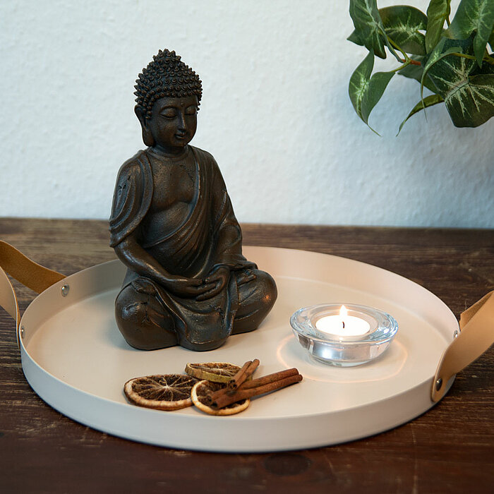 Auf einem Holztisch steht ein rundes Tablett mit Lederriemen, auf dem sich eine Buddha-Statue, eine brennende Teelichtkerze in einem Glashalter und getrocknete Orangenscheiben sowie Zimtstangen befinden. Rechts im Hintergrund ist eine grüne Pflanze in ein