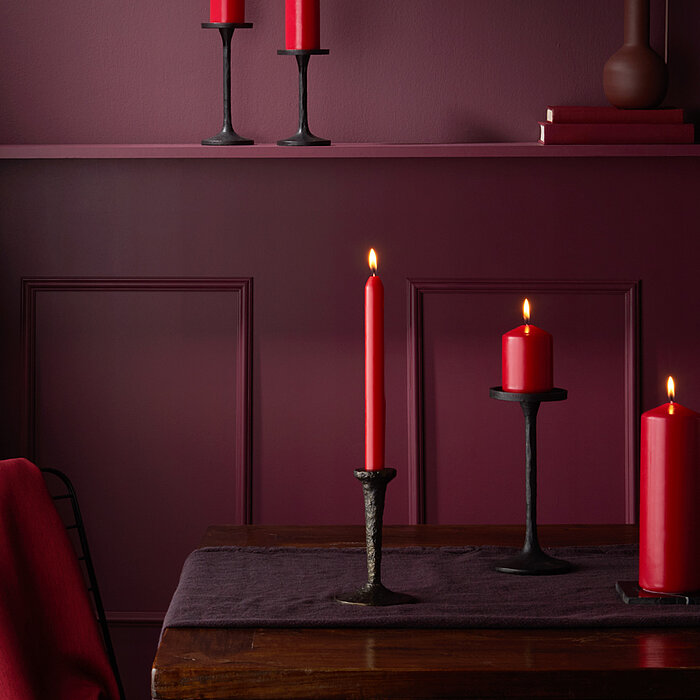Vier brennende Bolsius Haushaltskerzen in der Farbe Rot in verschiedenen Größen und Formen stehen auf einem dunkelbraunen Tisch. Die Kerzen sind auf schwarzen Kerzenhaltern platziert. Im Hintergrund ist eine weinrote Wand mit dekorativen Elementen und ein