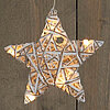 Ein leuchtender Stern aus weißem Rattan, der an einer Schnur hängt, vor einem holzgetäfelten Hintergrund. Die Lichter im Inneren des Sterns strahlen warmes Licht aus und wirken einladend und gemütlich. Der Stern ist mit kleinen Glitzerpartikeln dekoriert,