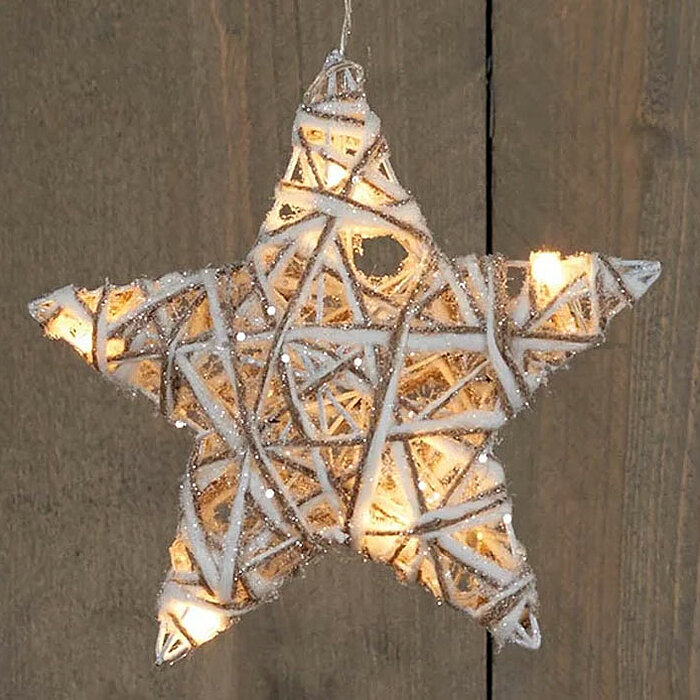 Ein leuchtender Stern aus weißem Rattan, der an einer Schnur hängt, vor einem holzgetäfelten Hintergrund. Die Lichter im Inneren des Sterns strahlen warmes Licht aus und wirken einladend und gemütlich. Der Stern ist mit kleinen Glitzerpartikeln dekoriert,