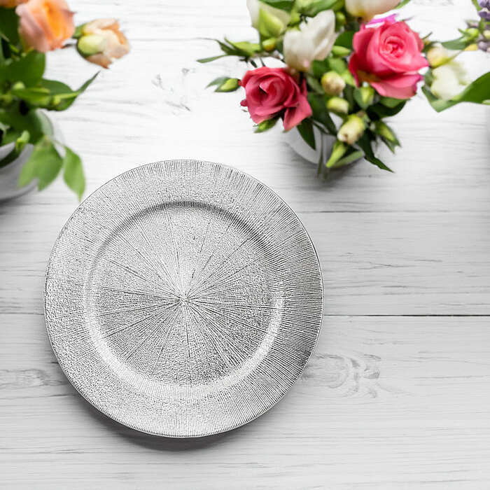 Ein silberner Teller liegt mittig auf einer weißen Holzfläche. Um den Teller herum sind vier Vasen mit frischen Blumen arrangiert, darunter Rosen in Pink und Weiß sowie andere bunte Blüten in Orange und Lila. Das Arrangement wirkt elegant und erfrischend.