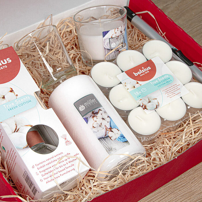 In einer roten Geschenkbox befinden sich eine Auswahl von DecoLite Produkten: ein Raumduft-Set mit weißem Diffuser und Duftstäbchen, eine große weiße Stumpenkerze, ein Paket mit weißen Stövchenkerzen und zwei klare Glasbehälter mit Duftkerzen. Im Hintergr