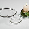 Eine brennende weiße Stumpenkerze, platziert auf einem runden, durchsichtigen Sandra Rich Kerzenteller, dekoriert mit grünem Moos und Efeu. Daneben liegen zwei leere Sandra Rich Glasteller in verschiedenen Größen auf einer weißen Oberfläche.