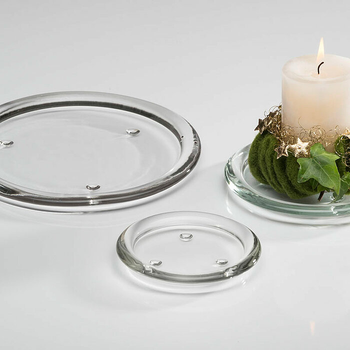 Eine brennende weiße Stumpenkerze, platziert auf einem runden, durchsichtigen Sandra Rich Kerzenteller, dekoriert mit grünem Moos und Efeu. Daneben liegen zwei leere Sandra Rich Glasteller in verschiedenen Größen auf einer weißen Oberfläche.