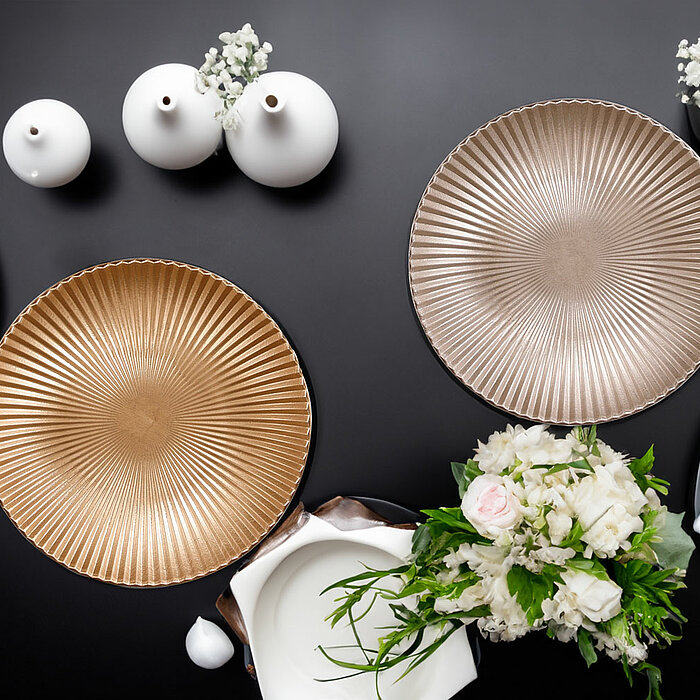 Zwei dekorative Teller vom Hersteller Edelman in Gold und Silber liegen auf einem schwarzen Tisch. Die Teller sind auf schwarzen Tellern platziert und mit weißen Stoffservietten dekoriert. Zusätzlich stehen weiße Vasen mit zarten weißen Blüten sowie weiße