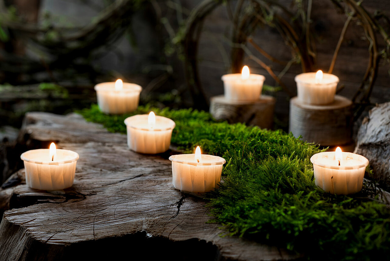 Mehrere brennende Stearin Kerzen in Form von Teelichtern im Papier-Cup auf einem mit Moos bewachsenen Baumstumpf.