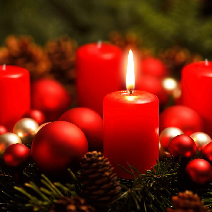 Fünf brennende rote Kerzen unterschiedlicher Größen stehen auf einem Adventskranz aus Tannenzweigen. Um die Kerzen herum sind rote und goldene Christbaumkugeln sowie Tannenzapfen verteilt. Die warme Flamme der Kerzen und das grüne Tannengrün schaffen eine