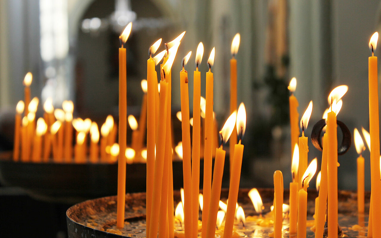 Auf dem Bild sieht man zwei Schalen mit jeweils einer Vielzahl von brennenden Opferkerzen in der Farbe natür. Sie haben unterschiedliche längen und erhellen das gesamte Bild. Im Hintergrund sieht man dass sich die Szene im inneren einer Kirche stattfindet.