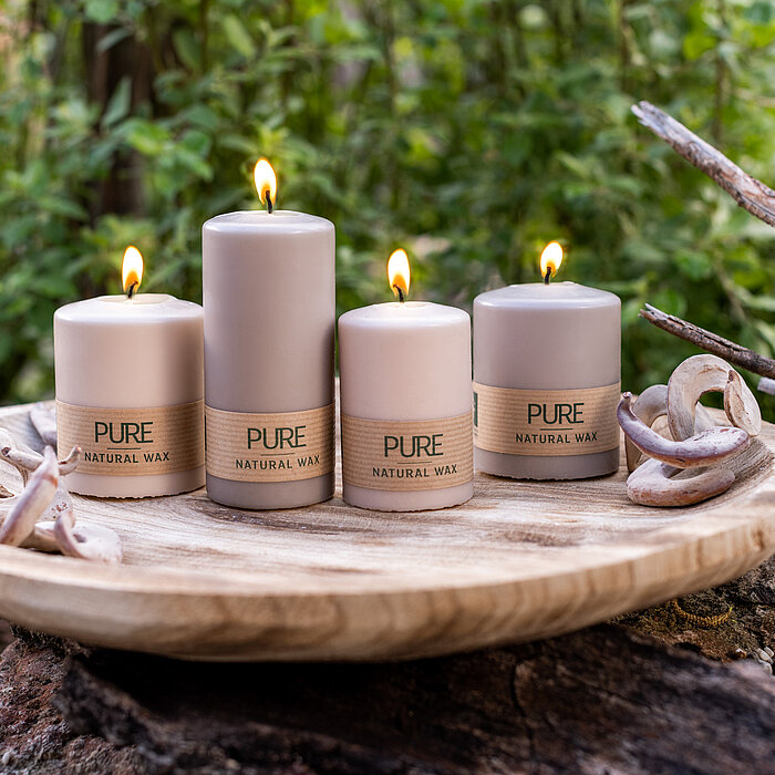 Vier brennende Kerzen des Herstellers Wenzel, in einer neutralen Beige-Farbe, stehen auf einem runden Holztablett. Die Kerzen haben verschiedene Größen und tragen die Aufschrift "PURE Natural Wax". Das Arrangement steht auf einem Baumstamm oder einer Stei