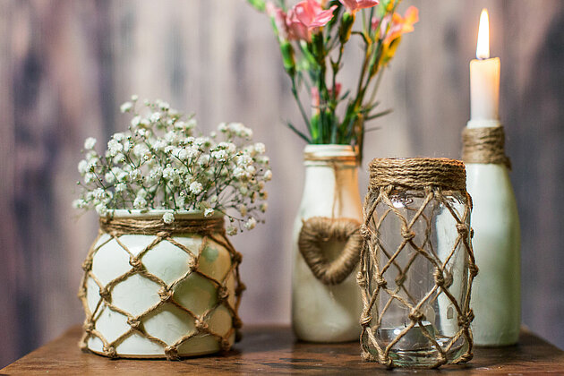 Auf einem rustikalen Holztisch stehen verschiedene Glas- und Keramikvasen. Zwei der Vasen sind liebevoll mit einem Makramee-Netz umspannt. Im Hintergrund sieht man zwei weitere Vasen, die mit Makramee-Kordeln kunstvoll verziert sind. In den Vasen stecken bunte Blumen und eine elegante Stabkerze. Das gesamte Bild verströmt eine warme, rustikale Ruhe und erinnert an die einfachen, schönen Dinge des Lebens.