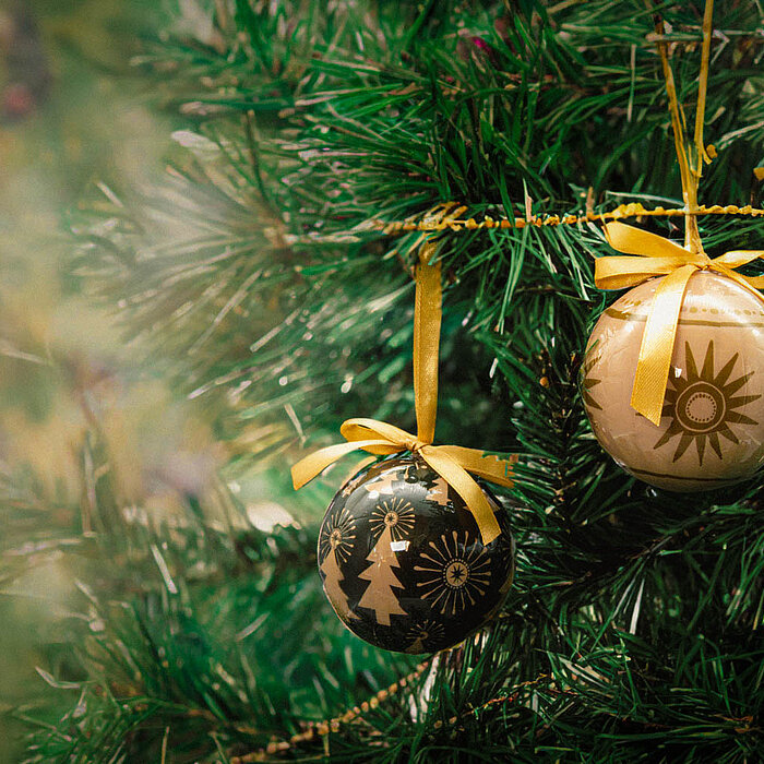 Zwei festliche Kugelornamente hängen an einem üppigen grünen Weihnachtsbaum. Eines ist schwarz mit goldenen Tannenbaum- und Sternmotiven, das andere goldfarben mit einem großen Sonnenmotiv. Beide Kugeln sind mit goldfarbenen Schleifen verziert und strahle