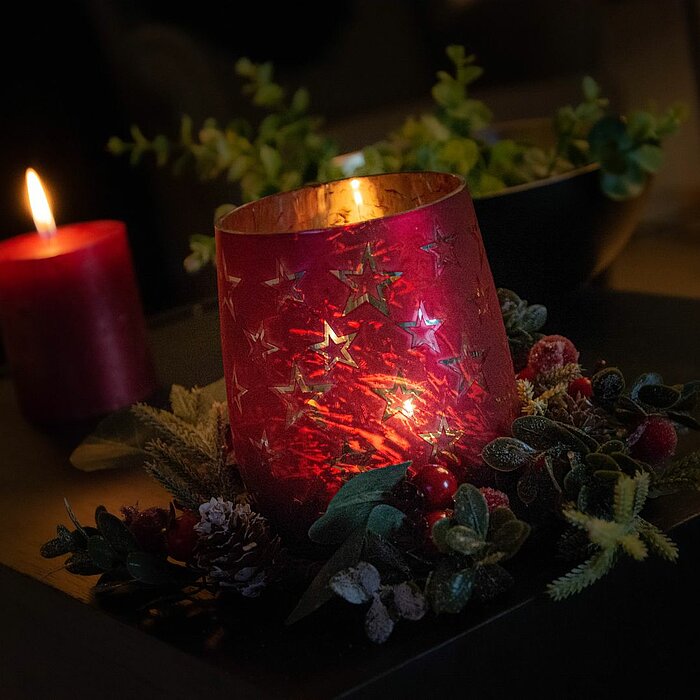Eine brennende, rote Stumpenkerze steht auf einem dunklen Tisch. Die Kerze ist von einem Kranz aus grünen Zweigen, kleinen Tannenzapfen und roten Beeren umgeben. Links im Bild ist eine grüne Pflanze in einem goldenen Blumentopf zu sehen.
