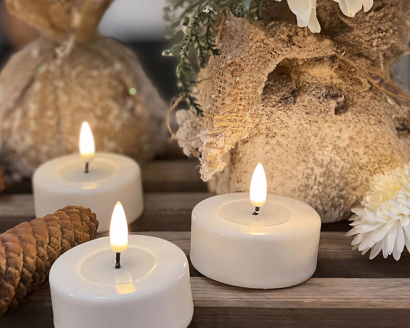Leuchtende LED Teelichter stehen auf einer groben Holzbank, neben einem Tannenzapfen und weißen Blüten, die in Leinensäcke gepflanzt wurden.