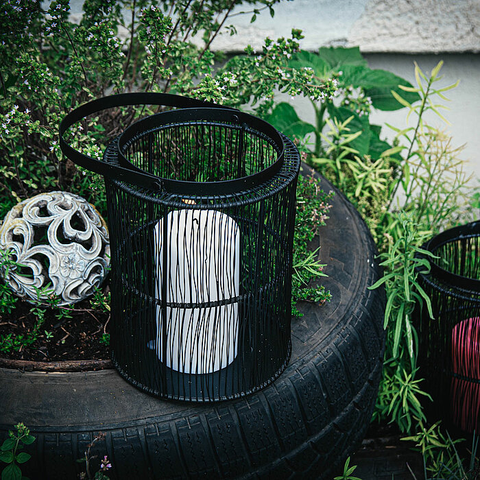 Eine brennende Stumpenkerze steht in einem schwarzen, drahtgeflechtartigen Kerzenhalter auf einem weißen Möbelstück. Die Wand hinter der Kerze ist mit einem grauen, floralen Tapetenmuster verziert. Die Beleuchtung der Kerze schafft eine warme und gemütlic