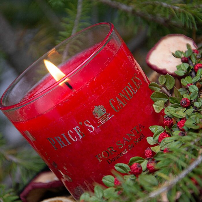 Eine brennende rote Kerze von Price's Candles in einem Glasbehälter, auf dem der Schriftzug "Price's Candles" zu lesen ist. Die Kerze ist umgeben von Tannenzweigen und kleinen roten Beeren, die die festliche Stimmung der Weihnachtszeit unterstreichen.