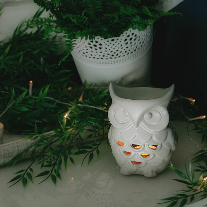 Ein weißer Keramik-Eulen-Dekorationsgegenstand befindet sich auf einem weißen Tisch. Die Eule hat leuchtende Ausschnitte, die eine warme orangefarbene Beleuchtung ausstrahlen. Um den Tisch herum sind grüne Zweige und ein Topf mit Farn angeordnet. Kleine L