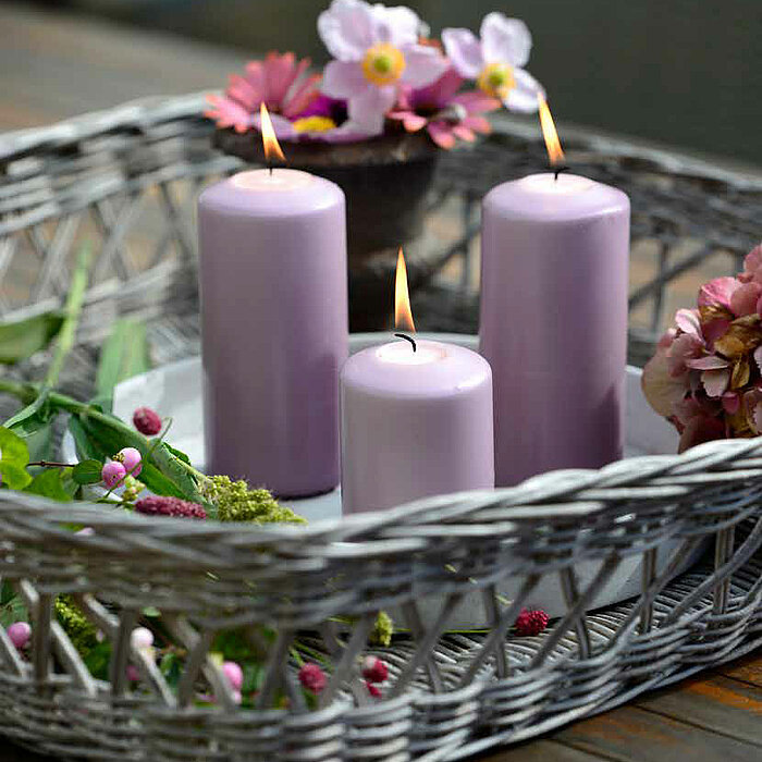 Drei brennende Müller-Stumpenkerzen in hellen Fliederfarben, die auf einem Tablett in einem grauen geflochtenen Korb aufgestellt sind. Umgeben sind die Kerzen von grünen Pflanzen, pinken und lila Blumen. Im Hintergrund sieht man einen Holztisch.