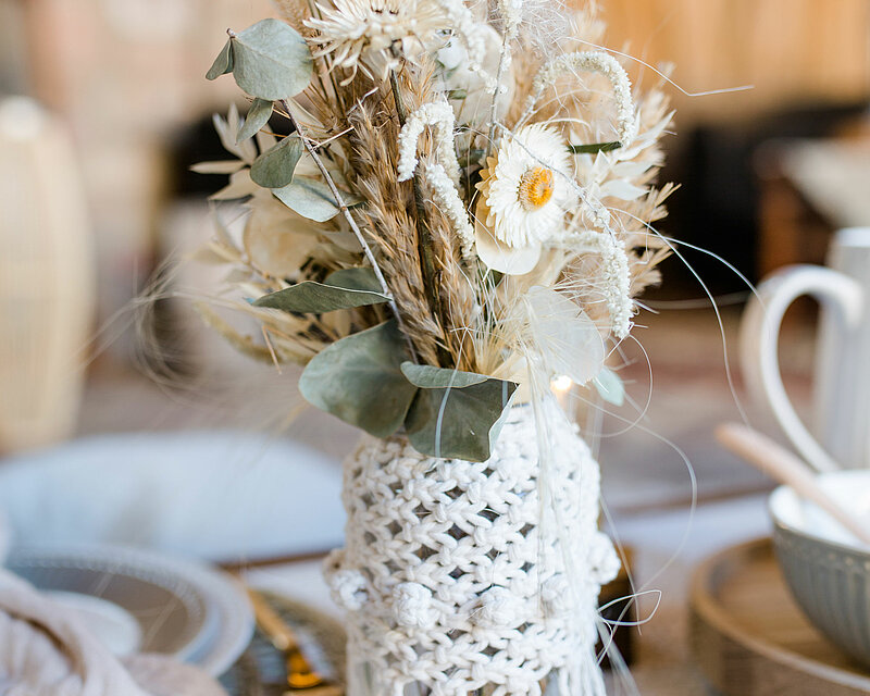 Auf einem romantisch und hell eingedeckten Tisch steht eine transparente Glasflasche, die mit einem weißen Makramee-Netz und kunstvollen Knoten umspannt ist. In der Flasche befinden sich verschiedene getrocknete Blumen und Gräser. Das gesamte Bild verströmt eine ländliche, rustikale Ausstrahlung und lädt zum Verweilen ein.