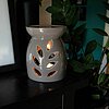Ein brennender Duftölbrenner in Form einer grauen Vase mit ausgeschnittenen Blattmotiven, durch die das Licht scheint. Der Brenner steht auf einem dunklen Holzregal, neben einer Schwarz-Weiß-Fotografie im Hintergrund. Vor dem Brenner sind grüne Blätter ei