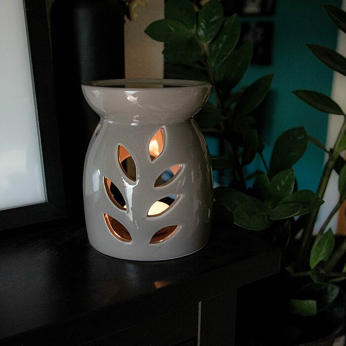 Ein brennender Duftölbrenner in Form einer grauen Vase mit ausgeschnittenen Blattmotiven, durch die das Licht scheint. Der Brenner steht auf einem dunklen Holzregal, neben einer Schwarz-Weiß-Fotografie im Hintergrund. Vor dem Brenner sind grüne Blätter ei