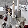 Drei brennende weiße Kerzen des Herstellers Müller in grauen, zylinderförmigen Kerzenhaltern aus Beton. Die Kerzen stehen auf einem weißen Tablett, das mit silbernen und roten Kugeln sowie Tannenzapfen dekoriert ist. Ein silbernes Rentier und ein glitzern