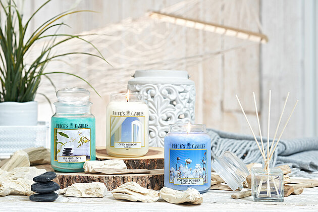 In einem hellen und maritim eingerichteten Zimmer stehen auf einem weißen Holztisch mehrere rustikale Holzscheiben. Auf den Holzscheiben stehen jeweils ein großes Apothekerglas von Pricers mit den Düften "Spa Moments", "Open Window" und "Cotton Powder". Rechts daneben steht ein Raumduft-Diffuser. Um die Gläser herum sind einige maritime Gegenstände verteilt, wie Treibholz und meerverwaschene Steine. Im Hintergrund sieht man leicht verschwommen eine Hängematte aus handgeknüpftem Stoff. Die Szene strahlt eine entspannte und küstennahe Atmosphäre aus, die zum Träumen und Verweilen einlädt.